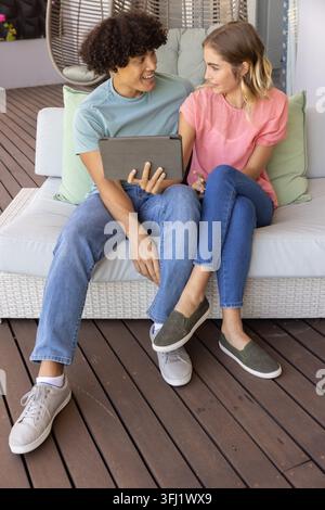 Couple sitting on white wicker sofa on wooden deck, wearing casual tees and jeans, sharing tablet. Patio, outdoors, conversation, device, relaxation, Stock Photo