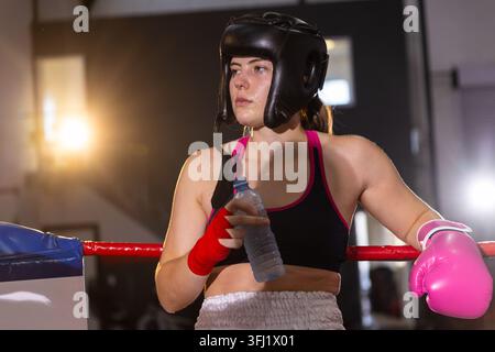 Female boxer leaning on red rope in ring corner, displaying headgear and pink glove, holding bottle. Boxing, athlete, gym, train Stock Photo