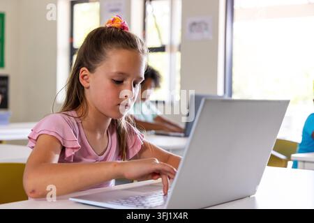 Classmates with girl typing on silver laptop in class wearing pink ruffle top and scrunchie. Classroom, student, education, learning, computer, daylig Stock Photo