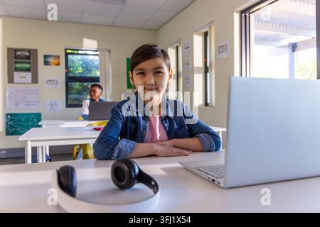 Asian girl and African American boy children sitting in class with denim jacket laptop headphones. Education, learning, students, classroom, technolog Stock Photo