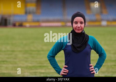 Confident Muslim woman athlete wearing a sports hijab and long-sleeve top stands on a grass stadium field with hands on hips, conveying determination, Stock Photo