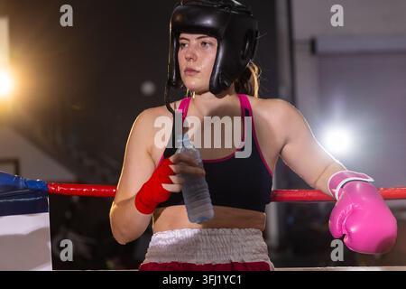 Female boxer pausing in boxing ring, leaning on red ropes, holding water bottle, wearing pink glove. Athlete, fighter, focus, en Stock Photo
