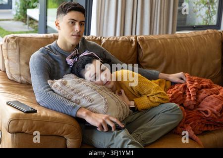 Couple sitting on sofa at home, woman in mustard sweater reclining on pillow, man holding remote. Cozy, lounge, knit, blanket, leather, greenery, smar Stock Photo
