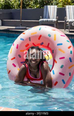 African American woman leaning through pink donut in pool, wearing red swimsuit and sunglasses. Inflatable, float, outdoor, back Stock Photo