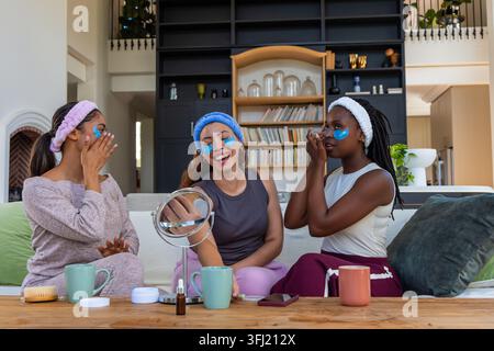 African American women friends sitting on sofa, wearing headbands and eye patches, holding mirror. Cozy, pampering, skincare, girlfriends, selfcare, h Stock Photo