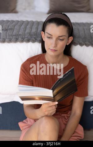Mid-adult female sitting on bedroom floor leaning by bed reading paperback, in rust top, headband. Cozy, home, book, landscape, soft, natural, calm Stock Photo