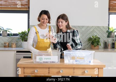 Diverse female friends in overalls and plaid shirt sorting at kitchen island PAPER and PLASTIC bins. Teamwork, recycling, sustainable, daylight, brigh Stock Photo