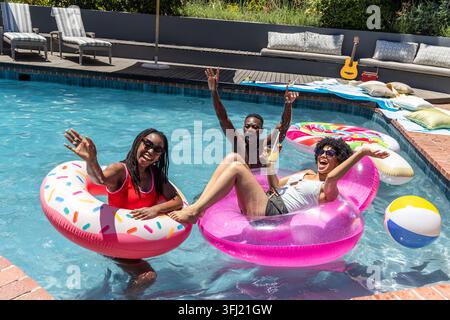 African American friends floating on sprinkle donut and pink ring in backyard pool, holding cup. Outdoor, summer, relaxation, gr Stock Photo