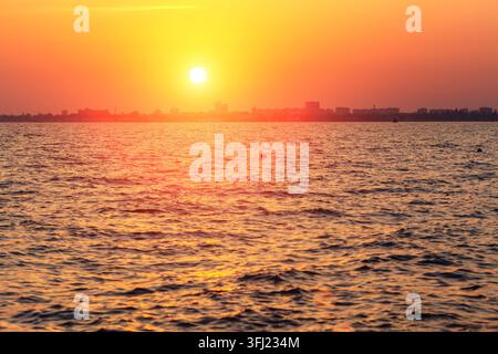 Vivid golden sunset over a calm sea with the glowing sun reflecting on rippling water and a distant city skyline silhouette on the horizon at dusk Stock Photo