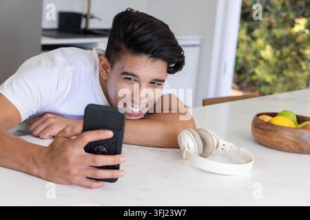 Leaning on marble countertop in kitchen, African American man holding black smartphone, smiling. Lifestyle, home, modern, casual Stock Photo
