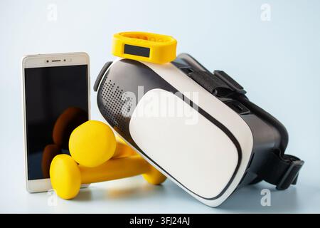 Composition of gadgets for tracking healthy training on blue background. Set of VR headset with smartwatch and mobile phone with  yellow dumbbells. Stock Photo