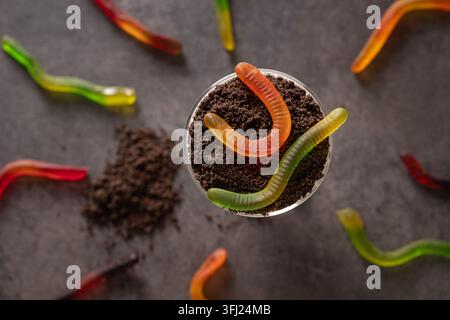 Small Halloween Dirt cake topped with chocolate cookie crumbs and colorful gummy worms, surrounded by candy on dark surface. Fun spooky party food. Stock Photo