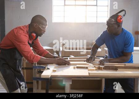Two carpenters are aligning and measuring wooden boards on wooden workbench inside woodworking shop. Craftsmanship, woodworking, Stock Photo