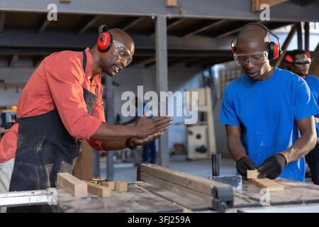 African American male carpenters feeding wooden plank into table saw, wearing ear muffs at workshop. Craftsmanship, industrial, Stock Photo