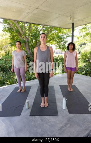 Three women doing yoga on mats under concrete overhang in workout gear, bottles, center with watch. Outdoor, fitness, group, ses Stock Photo