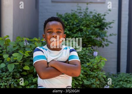 African American boy in striped tee smiling with arms crossed standing in courtyard by shrubs. Child, outdoor, garden, greenery, brick, daylight, frie Stock Photo