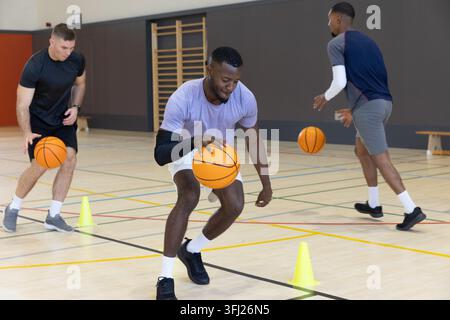 Diverse male teammates performing dribbling drill on gym court with orange basketballs and cones. Athlete, training, teamwork, f Stock Photo