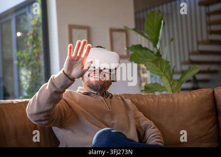Mature Asian man wearing white VR headset, reaching forward while sitting on brown leather sofa. Wearable, immersive, gadget, mo Stock Photo
