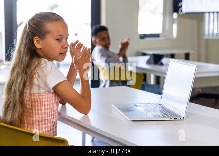 Diverse schoolkids girl in pink dress sitting in classroom clapping near silver laptop, copy space. Classmates, collaboration, l Stock Photo