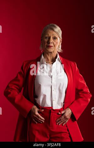 Senior woman posing in studio with red backdrop wearing red suit white shirt gold necklace Stock Photo