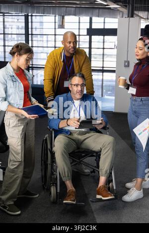 Diverse coworkers gathering around senior man in wheelchair, using tablet and charts at office. Teamwork, leadership, inclusion, accessibility, device Stock Photo
