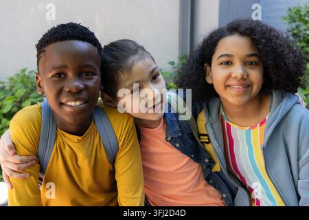 Diverse pre-teen friends sitting on bench in courtyard with backpacks, denim jacket, gray hoodie. Students, outdoor, schoolyard, colorful, friendship, Stock Photo
