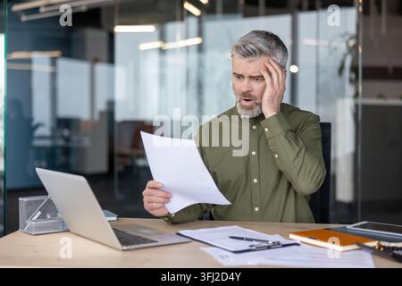 Business stress and financial crisis, mature businessman reading bad news on documents at office desk with laptop and feeling shocked and worried about company performance and economic loss. Stock Photo