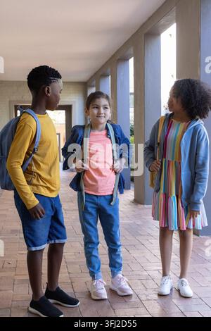 Diverse child classmates talking and standing in school walkway with backpacks, denim jacket. Students, peers, trio, corridor, columns, daylight, gree Stock Photo