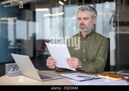 Businessman reviewing documents and financial analysis on a laptop at his office desk. Focused professional working on corporate strategy and reports to ensure accuracy and business data efficiency. Stock Photo