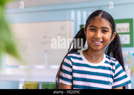 Indian child smiling, facing camera at school by whiteboard in striped tee, copy space. Classroom, student, portrait, cheerful, bright, plant, colorfu Stock Photo