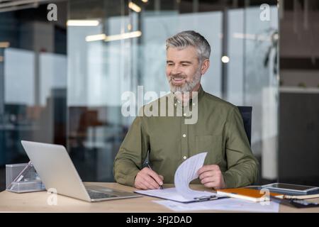 Businessman working on laptop and documents at desk in office. Professional man doing financial analysis and corporate planning for business growth while reviewing important company paperwork. Stock Photo