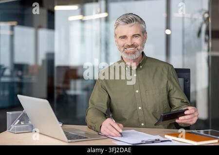 Businessman working at desk with laptop and documents, professional man using calculator for financial analysis in modern office environment. Experienced executive managing business strategy and data. Stock Photo