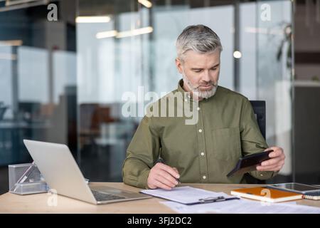 Financial analysis and business planning, mature businessman using a calculator and laptop for accounting tasks at a desk in a modern office space. Professional manager reviewing documents. Stock Photo