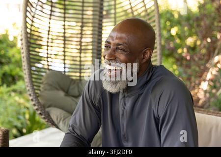 Senior African American man sitting, laughing in dark zip-up pullover on woven egg chair at patio. Porch, outdoor, garden, green Stock Photo