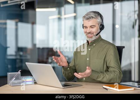 Remote work and virtual meeting, mature businessman using laptop and headset for video call at office desk to discuss business strategy and project with colleagues during a professional conference. Stock Photo