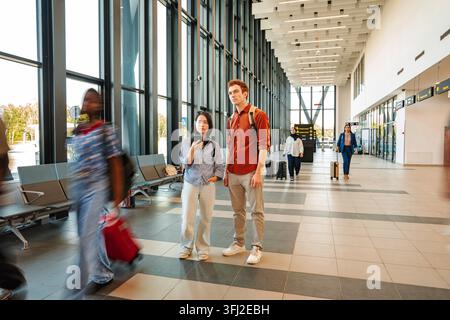 Young multicultural adult travelers standing in a modern airport terminal while other people are in motion blur Stock Photo