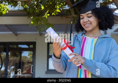 African American teen female in blue hoodie holding red ribbon diploma cap at entrance, copy space. Student, graduate, campus, o Stock Photo