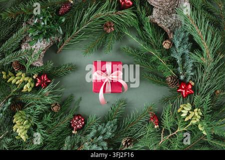 Overhead view of a wrapped Christmas gift surrounded by evergreen fir branches, thuja, pinecones and assorted festive red Christmas baubles and ornaments on a green background Stock Photo