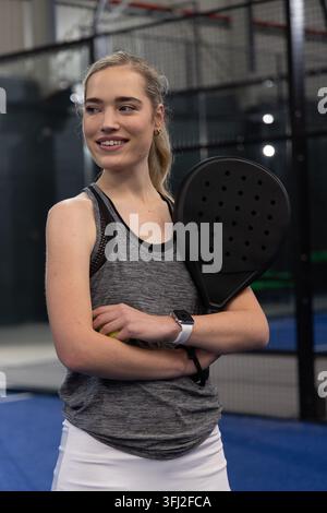 Female standing on padel court holding racket and ball, wearing grey tank and white skirt. Woman, athlete, racquet, activewear, Stock Photo