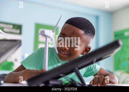 African American child boy leaning over school desk examining wind turbine model with black tablet. Student, classroom, renewable, energy, solar, scie Stock Photo