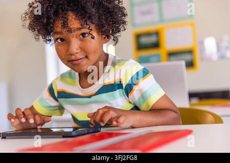 African American boy sitting at desk in classroom wearing striped tee using tablet near red folder. Student, device, education, learning, sunlit, scho Stock Photo