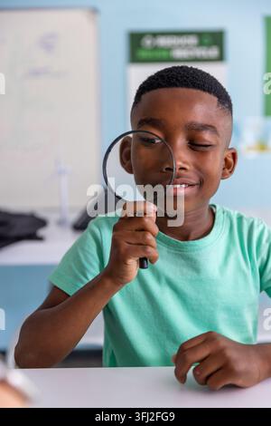 African American male child wearing mint tee holding magnifier, peering at desk in classroom. Student, curiosity, exploration, l Stock Photo