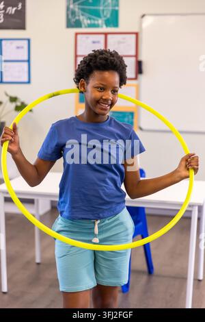 African American child wearing blue tee and aqua shorts holding yellow hula hoop in classroom. Kid, student, school, education, Stock Photo