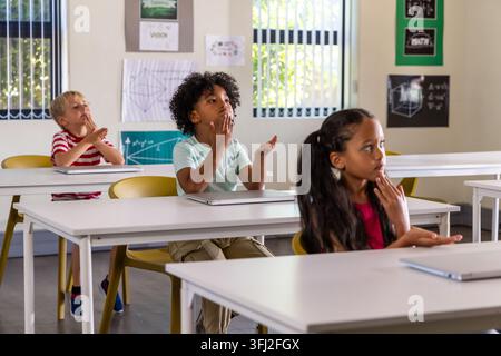 Diverse children classmates sitting at white desks in classroom gesturing near closed laptops. Learning, participation, focus, c Stock Photo