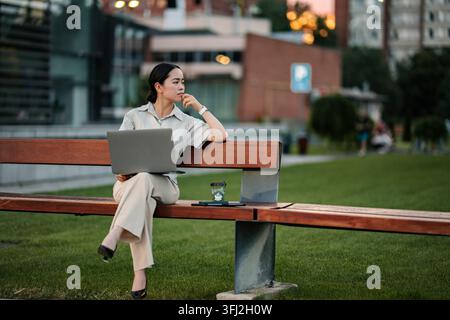 Young adult woman thinking while working on laptop in city park Stock Photo