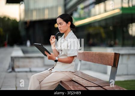 Young Asian woman thinking with digital tablet on urban bench Stock Photo