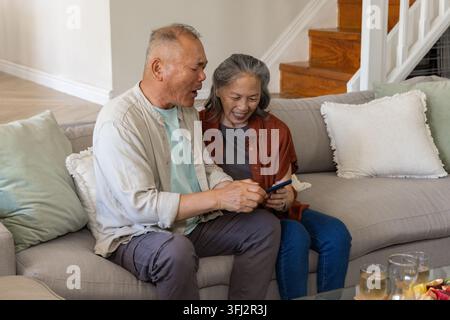 Senior Asian couple sitting on sofa in living room with green pillows, man holding smartphone. Cozy, warm, intimate, home, staircase, minimalist, tech Stock Photo