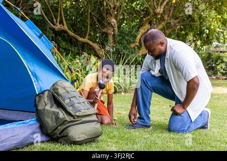 African American father and son kneeling, using black mallet to secure blue dome tent on lawn. Outdoors, camping, family, teamwork, nature, sunlit, ba Stock Photo