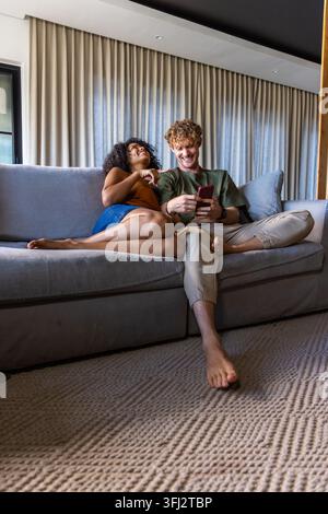 Diverse couple on sofa in living room, woman reclining in orange top, man holding red smartphone. Cozy, modern, relaxed, casual, Stock Photo