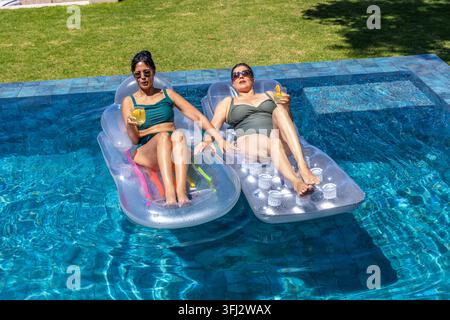 Diverse female friends reclining on clear floats, wearing sunglasses poolside with yellow drinks. Outdoor, vibrant, sunlit, rela Stock Photo